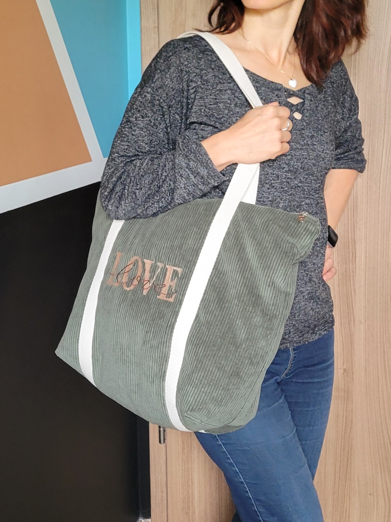 Grand sac cabas en velours côtelé couleur kaki, orné d'une broderie "LOVE" sur le devant. Il se ferme entièrement grâce à une fermeture éclair dorée, garantissant sécurité et style, et se porte sur l'épaule grâce à deux belles anses écrues.